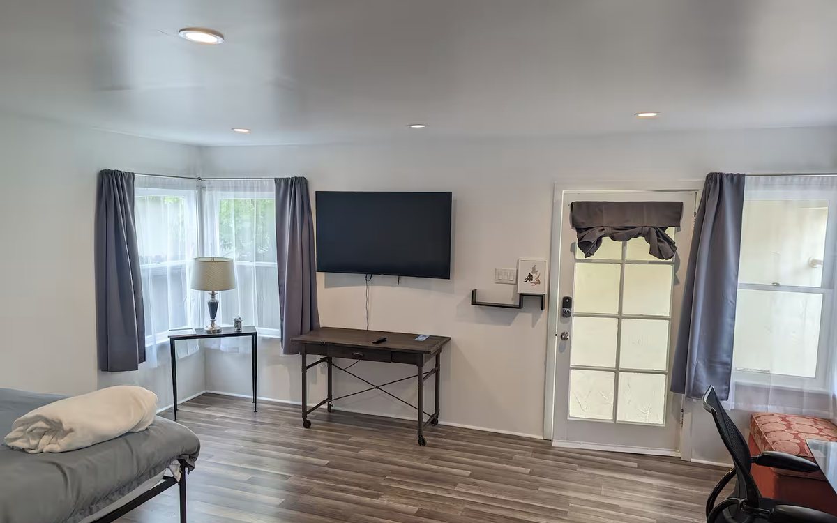 The main room: bed at left, wall-mounted television, a rolling standing desk, and the private entrance door. Blackout curtains at both windows.